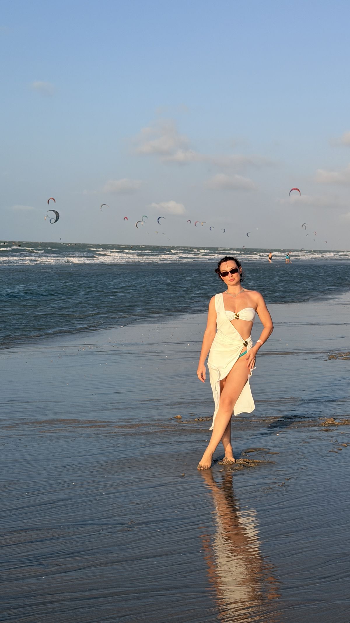 Lidiane Grigoletto caminhando na beira-mar da Praia do Preá, com os kites coloridos no céu ao fundo